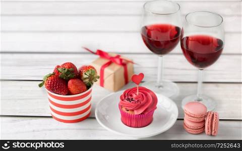 valentines day and sweets concept - frosted cupcake with red heart, macarons, strawberries, glasses of wine and gift box on grey wooden boards background. close up of red sweets for valentines day