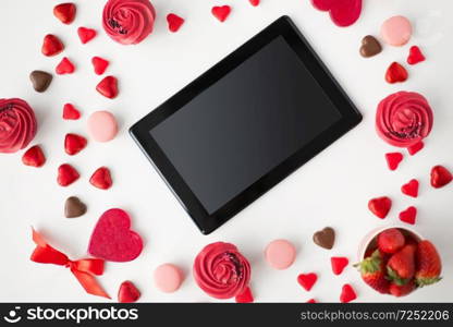 valentines day and technology concept - close up of tablet pc computer, frosted cupcakes, red heart shaped chocolate candies, macarons and strawberries. close up of tablet pc and sweets on valentines day