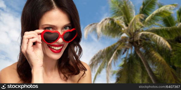 valentines day, beauty and people concept - close up of happy smiling young woman with red lipstick and heart shaped sunglasses over sky and palm tree background. woman in heart shaped shades over palm tree