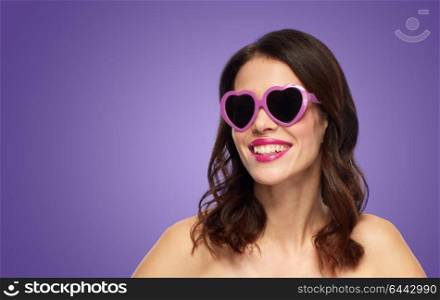 valentines day, beauty and people concept - close up of happy smiling young woman with berry lipstick and heart shaped sunglasses over ultra violet background. woman with heart shaped shades over ultra violet