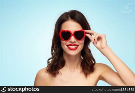 valentines day, beauty and people concept - happy smiling young woman with red lipstick and heart shaped sunglasses over blue background. woman with red lipstick and heart shaped shades. woman with red lipstick and heart shaped shades