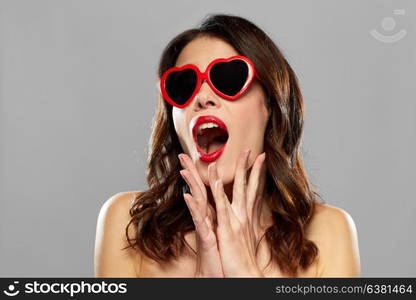 valentines day, beauty and people concept - happy smiling young woman with red lipstick and heart shaped sunglasses over gray background. woman with red lipstick and heart shaped shades