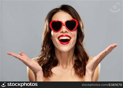 valentines day, beauty and people concept - happy smiling young woman with red lipstick and heart shaped sunglasses over gray background. woman with red lipstick and heart shaped shades