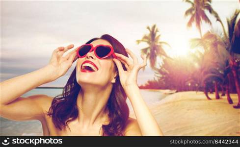 valentines day, beauty and people concept - happy smiling young woman with red lipstick and heart shaped sunglasses over exotic tropical beach with palm trees background. woman with red lipstick and heart shaped shades