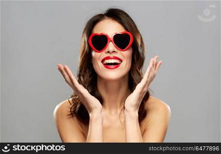 valentines day, beauty and people concept - happy smiling young woman with red lipstick and heart shaped sunglasses over gray background. woman with red lipstick and heart shaped shades. woman with red lipstick and heart shaped shades