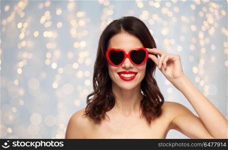 valentines day, beauty and people concept - happy smiling young woman with red lipstick and heart shaped sunglasses over holidays lights background. woman with red lipstick and heart shaped shades. woman with red lipstick and heart shaped shades