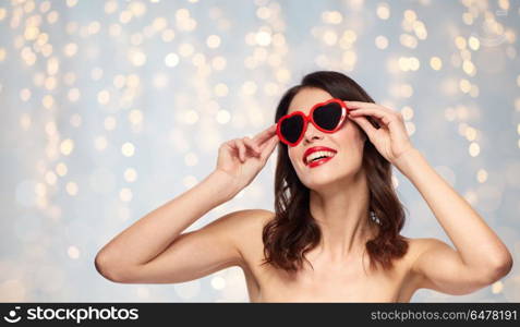 valentines day, beauty and people concept - happy smiling young woman with red lipstick and heart shaped sunglasses over holidays lights background. woman with red lipstick and heart shaped shades. woman with red lipstick and heart shaped shades