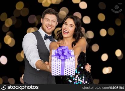 valentines day, birthday, anniversary and holidays concept - happy couple with gift box at party over lights on black background. happy couple with gift box at birthday party
