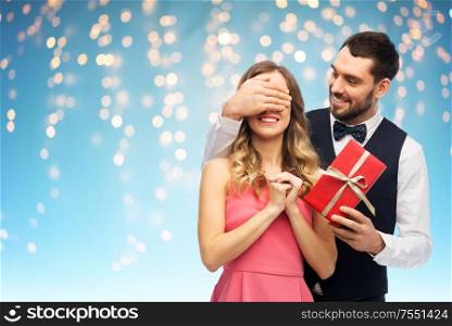 valentines day, couple, relationships and people concept - happy man giving woman surprise present over holiday lights on blue background. happy man giving woman surprise present