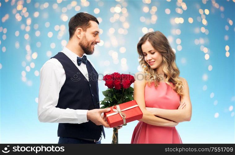 valentines day, couple, relationships and people concept - happy man giving woman flowers and present over holiday lights on blue background. happy man giving woman flowers and present