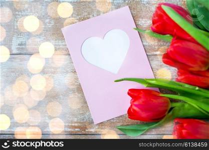 valentines day, greeting, love and holidays concept - close up of red tulips and greeting card with heart over lights