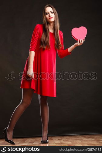 Valentines day happiness and relationships concept. Brunette woman long hair girl in red dress holding heart shaped gift box love symbol dark gray background.
