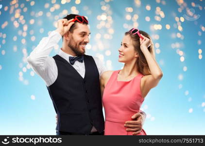 valentines day, love and people concept - happy couple in heart-shaped sunglasses over holiday lights on blue background. happy couple in heart-shaped sunglasses