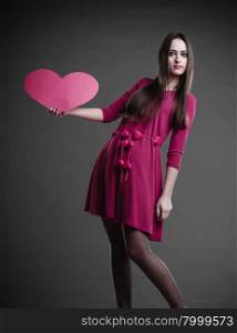 Valentines day love and relationships concept. Brunette long hair young woman in fuchsia dress holding heart love symbol dark gray background