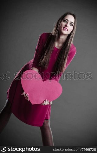 Valentines day love and relationships concept. Brunette long hair young woman in fuchsia dress holding heart love symbol dark gray background