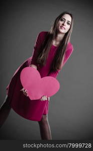 Valentines day love and relationships concept. Brunette long hair young woman in fuchsia dress holding heart love symbol dark gray background