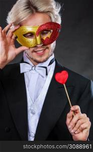 Valentines day. Love concept. Holidays and celebration. Elegant young guy wearing suit white shirt bow tie with carnival mask and heart stick in hand, on dark.. Man in carnival mask with heart stick love symbol