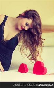 Valentines Day, love, feelings. Attractive seductive long haired woman in black dress lingerie on bed with red heart box gift. Indoor.