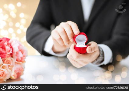 valentines day, proposal and engagement concept - close up of man with diamond ring in little red gift box and flowers on table over beige background with festive lights. man with diamond engagement ring in red gift box