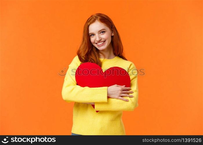 Valentines day, romance or relationship concept. Tender, lovely redhead girl waiting for her loved one, receive valentine card, hugging large heart sign and smiling happy, standing orange background.. Valentines day, romance or relationship concept. Tender, lovely redhead girl waiting for her loved one, receive valentine card, hugging large heart sign and smiling happy, standing orange background