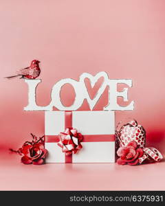 Valentines day romantic still life composition with word Love and greeting setting: gift box, ribbon, and decoration at red background, front view.
