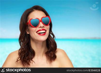 valentines day, summer vacation, travel and tourism concept - happy smiling young woman with red lipstick and heart shaped sunglasses over blue sea and sky background. woman with heart shaped shades over sea and sky