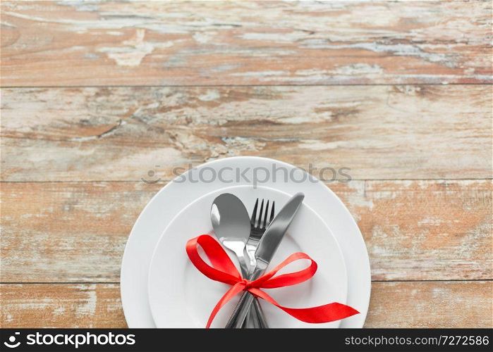 valentines day, table setting and festive dinner concept - plate with spoon, knife and fork tied with red ribbon on wooden background from top. cutlery tied with red ribbon on set of plates