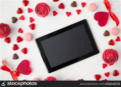 valentines day, technology and romantic concept - close up of tablet pc computer, frosted cupcakes, red heart shaped chocolate candies and macarons. close up of tablet pc and sweets on valentines day