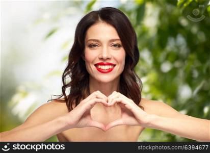 valentines day, wellness, organic and people concept - happy smiling young woman with red lipstick making hand heart gesture over green natural background. beautiful woman with red lipstick and hand heart