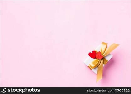 Valentines&rsquo; day background concept. White gift box with a golden bow ribbon and wood red hearts composition greeting card for happy love isolated on pink background with copy space. View from above