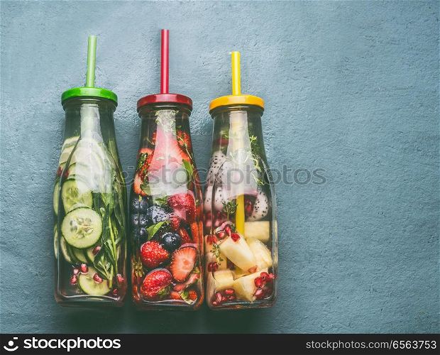 Variety of colorful infused water in bottles with fruits berries, cucumber, herbs and drink straws on gray background, top view. Tasty summer clean beverages for healthy lifestyle and fitness
