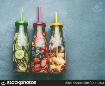 Variety of colorful infused water in bottles with fruits berries, cucumber, herbs and drink straws on gray background, top view. Tasty summer clean beverages for healthy lifestyle and fitness