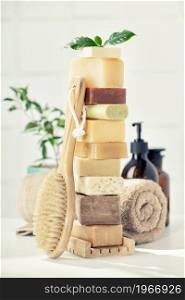 Variety of natural organic handmade soap bars on white bathroom countertop, wooden massage brush, towel and scincare accessories