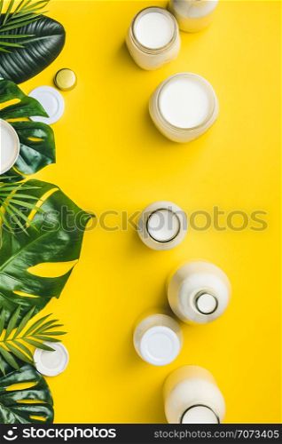 Various bottles of milk and tropical leaves on yellow background, flat lay. Vegan, vegetarian or clean eating concept