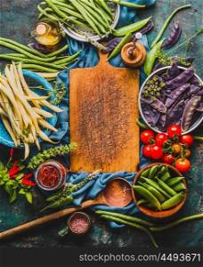 Various colorful pea and beans pods around empty cutting board with cooking ingredients and wooden spoon, top view, frame