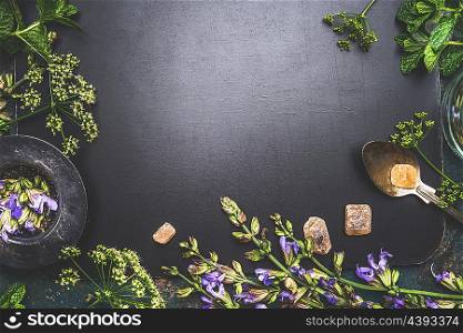 Various fresh healing herbs and flowers , tea strainer , spoon and sweets on dark background, top view, frame