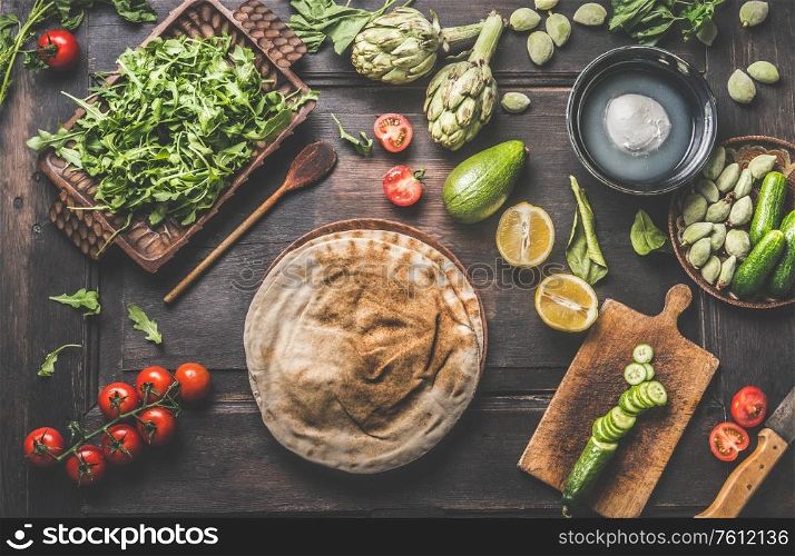Various fresh ingredients around tortilla or flatbread for tasty vegan wraps making. Fresh salad ingredients: avocado, lemon, arugula, tomatoes, green almonds, artichokes on wooden background