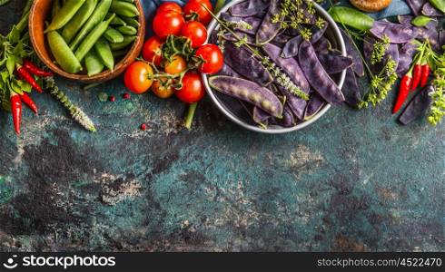 Various green and purple pea pods in bowls with cooking ingredients on dark rustic background, top view