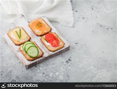Various healthy crackers with salmon and cheese, tomato and cucumber on wooden chopping board on light table background. Top view