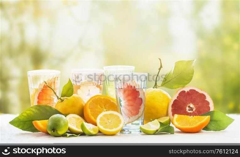 Various healthy summer lemonade in glasses with variety of organic citrus fruits: lime, oranges, grapefruit and lemon on table at green sunny garden nature background. Infused water. Copy space