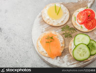 Various round healthy crackers with salmon and cheese, tomato and cucumber on wooden chopping board on light table background. Macro