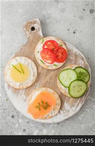 Various round healthy crackers with salmon and cheese, tomato and cucumber on wooden chopping board on light table background. Top view