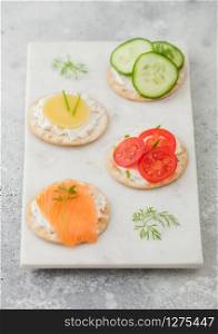 Various round healthy crackers with salmon and cheese, tomato and cucumber on marble board on light table background. Top view