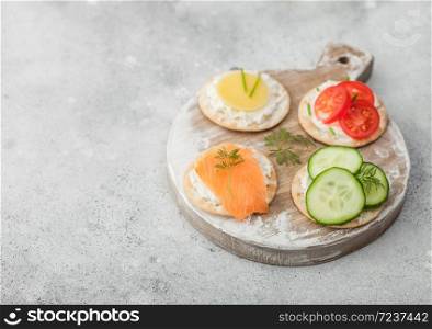 Various round healthy crackers with salmon and cheese, tomato and cucumber on wooden chopping board on light table background. Top view