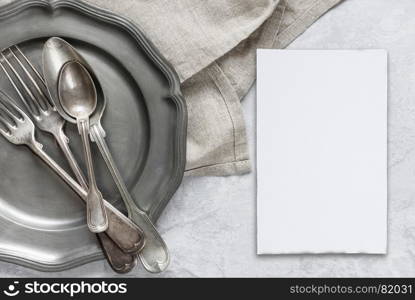 Various silverware on a pewter plate, gray flax napkin and white blanc paper card are on the background of gray concrete surface, with copy-space