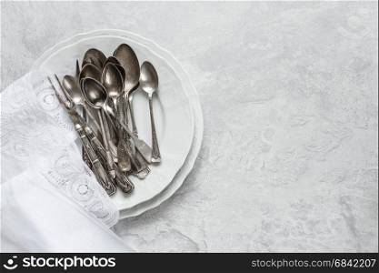 Various silverware on a porcelain plate and white napkin with Belgian lace are on the background of gray concrete surface, with copy-space