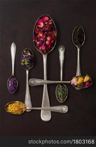 Various spoons with black and green tea, rose buds mix and blue mallow flowers and petals on brown background. Top view