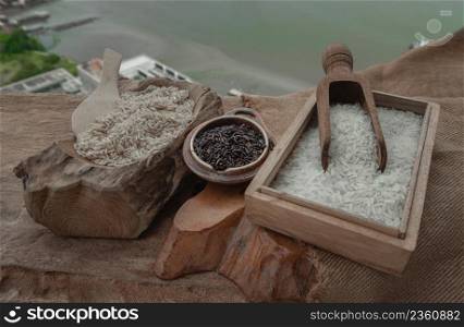 Various types of rice : Brown rice, Jasmine rice and Riceberry on old wooden table with river scenery. Organic raw rice collection, Healthy food and diet concept, Selective focus.