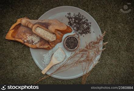 Various types of rice : Brown rice, Jasmine rice, Riceberry and dried flowers put on ceramic plate over green grass. Organic raw rice collection, Healthy food and diet concept, Overhead view, Selective focus.