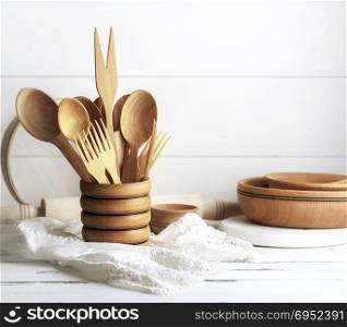 various wooden objects in a wooden jar, spoons, forks, rolling pin, plates and a sieve on a white wooden table